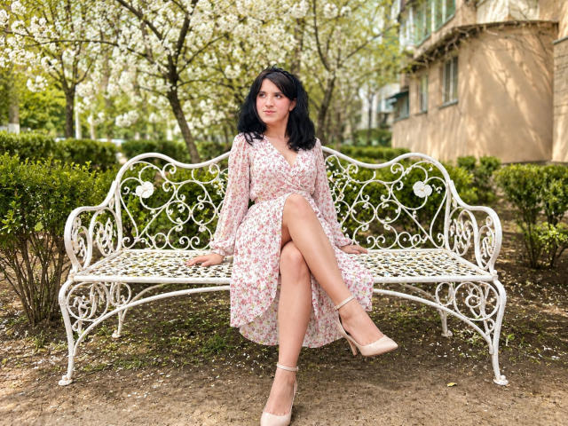 AnnaKimburli, avec des cheveux noirs, une robe à fleurs, les jambes croisées, un banc blanc orné, des talons beiges, assise dans un parc, avec un sourire doux, une poitrine de taille moyenne, une peau claire, un arrière-plan printanier..