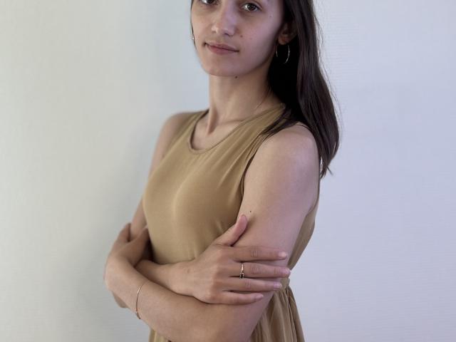 Joanna Vanderheide, con cabello castaño, vestida con un vestido beige sin mangas, brazos cruzados, pechos pequeños, sonrisa sutil, aretes de aro dorados, anillo en la mano izquierda, piel clara, maquillaje mínimo..