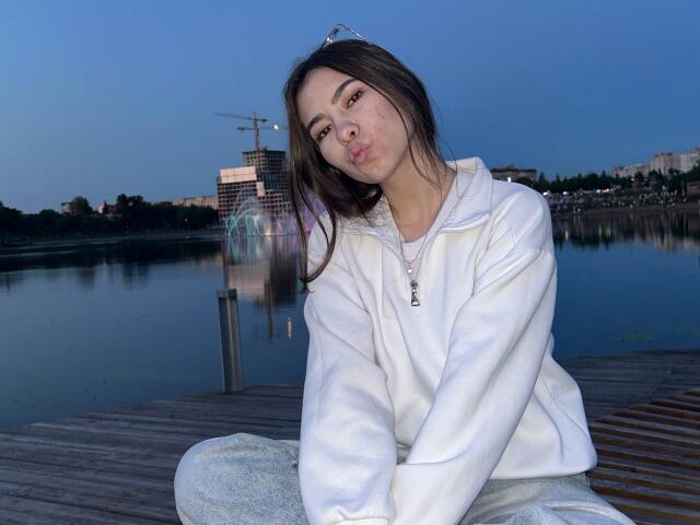 EmeraldCrivaro with pouty lips, wearing a white hoodie and grey pants, sitting on a wooden dock at dusk, reflecting in calm water, blue sky in background.