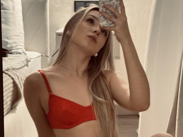 DanikaPlassmann with long blonde hair, red bra, taking a mirror selfie, showing her tits, holding a phone with a floral case, slightly parted lips, in a bedroom.