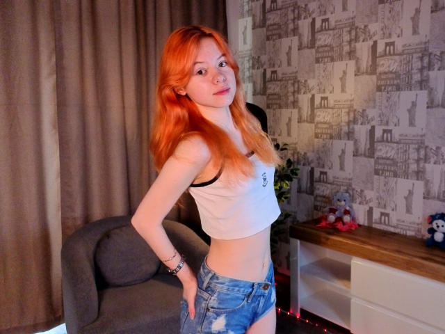 JoiMazey has long red hair, a white crop top, and blue distressed jeans, standing with a hand on her hip, showing her flat stomach and small ass.