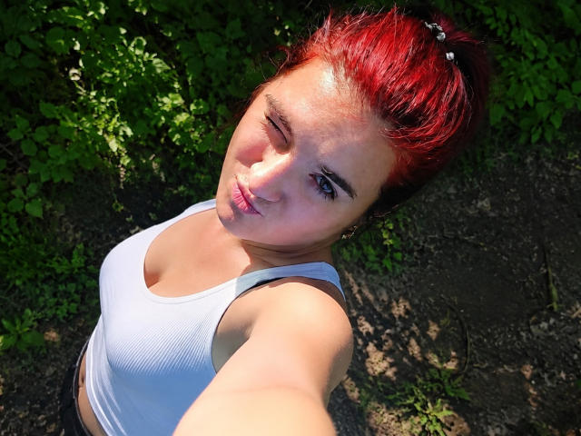 BrunetteBabe69 avec de grandes seins, vêtue d'une robe rouge en filet de pêche, lèvres boudeuses, longs cheveux noirs, boucles d'oreilles dorées, assise sur un lit avec un oreiller et un sac, dans une chambre..