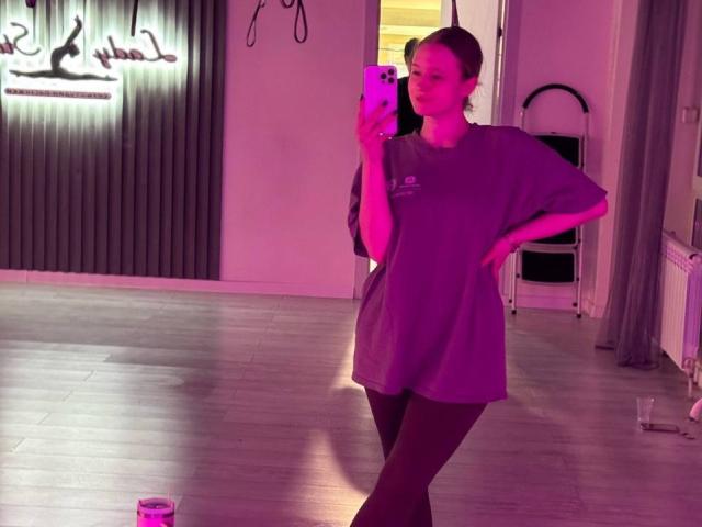 Shonna Sjolund em um quarto com iluminação roxa, vestindo uma camisa grande e collant preto, segurando um telefone, sorrindo, com a mão na cintura, em pé em um piso de madeira, reflexo em um espelho..