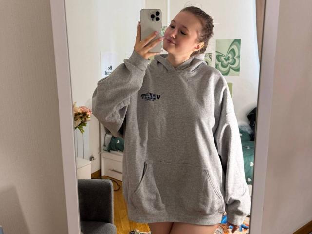 Shonna Sjolund, portant un sweat à capuche gris, prend un selfie devant un miroir avec son téléphone, souriant légèrement, avec des cheveux blonds attachés en un chignon désordonné, debout dans une pièce avec des planchers en bois..