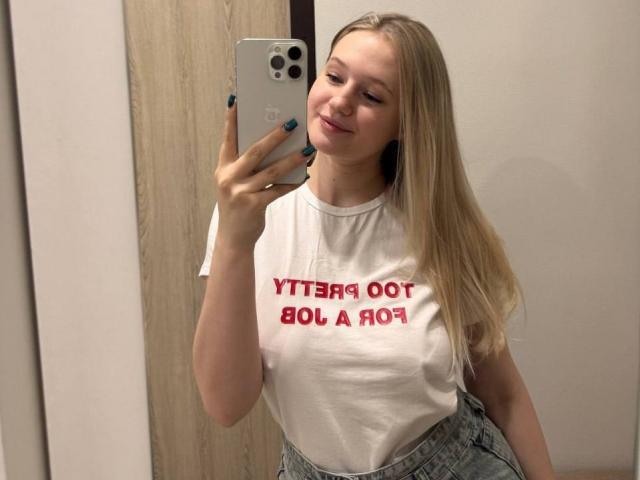 Shonna Sjolund, avec de longs cheveux blonds, souriante, portant une chemise blanche avec du texte rouge, tenant un iPhone argenté, autoportrait pris dans un miroir, portant un jean gris taille haute, vernis à ongles bleu turquoise..