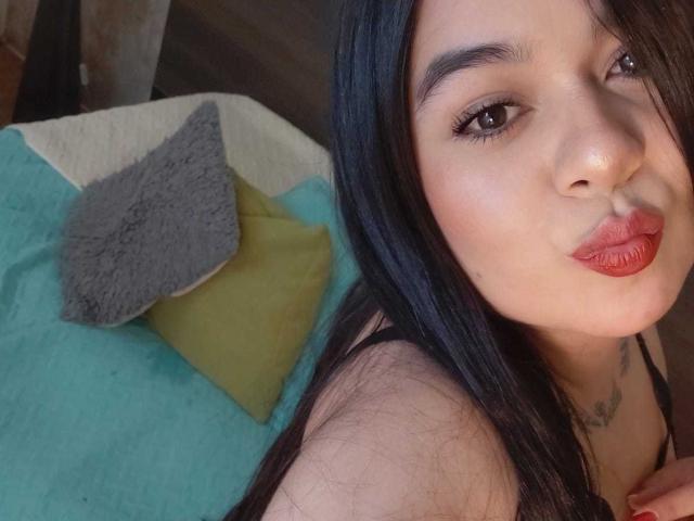 EveWillis with pouty red lips, black hair, and subtle makeup, wearing a black top, her small tits barely visible, lying on a bed with green and gray pillows.