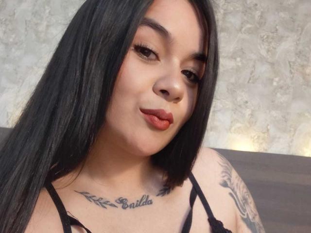 EveWillis with long black hair, brown eyes, and red lipstick, pouting slightly. She has tattoos on her chest and arms, wearing a black strappy top.