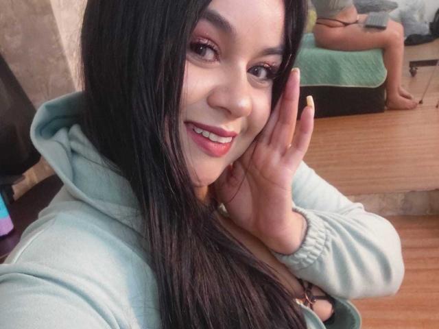 EveWillis with long black hair, smiling, wearing a green hoodie, touching her cheek, showing yellow nails, with a mirror reflecting her bare ass in the background.