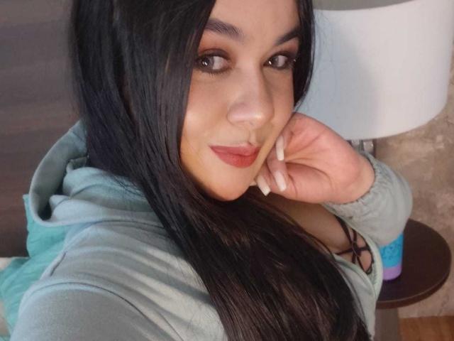 EveWillis with long black hair, wearing a green hoodie, has her hand near her face, looking at the camera with red lipstick and a slight smile. Her tits are visible.