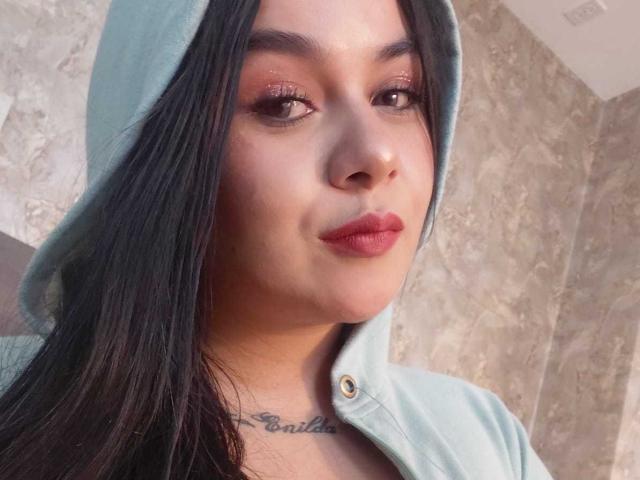 EveWillis with long black hair, wearing a light blue hoodie, has shiny pink lipstick and a tattoo below her neck. Her brown eyes are slightly open, looking forward.