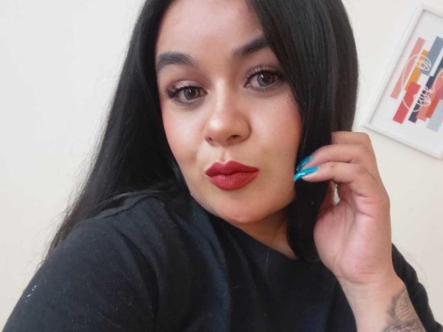 EveWillis has long black hair, red lipstick, and blue nails, with a tattoo on her right hand. She's wearing a black shirt, looking at the camera with a pout.