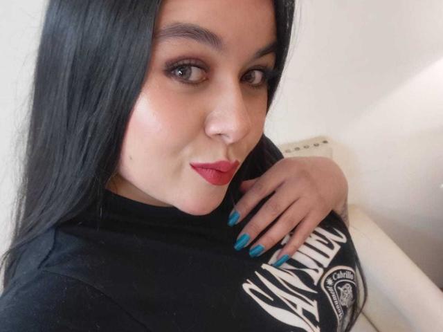EveWillis has long black hair, red lipstick, and blue nail polish. She's wearing a black shirt, with her hand on her chest, showing her tits. She's looking at the camera.