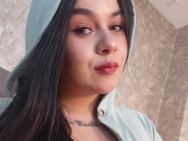 EveWillis has long black hair, wearing a light blue hoodie with a buttoned neck, subtle makeup with pink lipstick, and a tattoo under her chin. She looks slightly to the side.