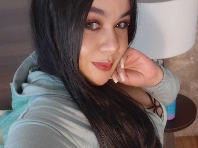 EveWillis has long black hair, brown eyes, and red lipstick. She's wearing a teal hoodie, with her hand near her face and a slight smile. Her tits are partially visible.
