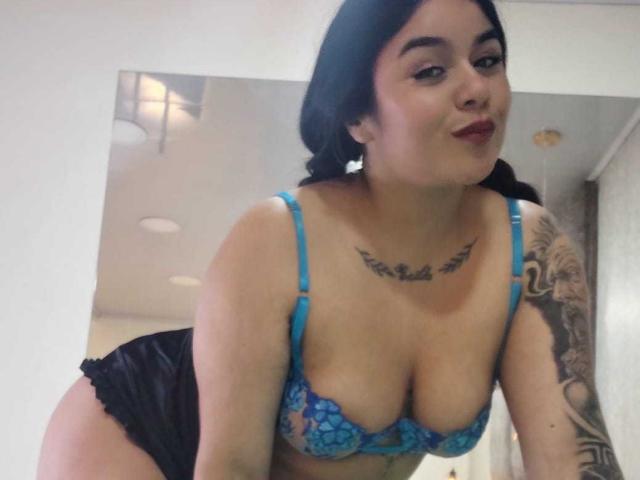EveWillis with dark hair, blue lace bra, black panties, tattoo on chest, tattoo on arm, leaning forward, smiling, showing her large tits, small ass, in a selfie.