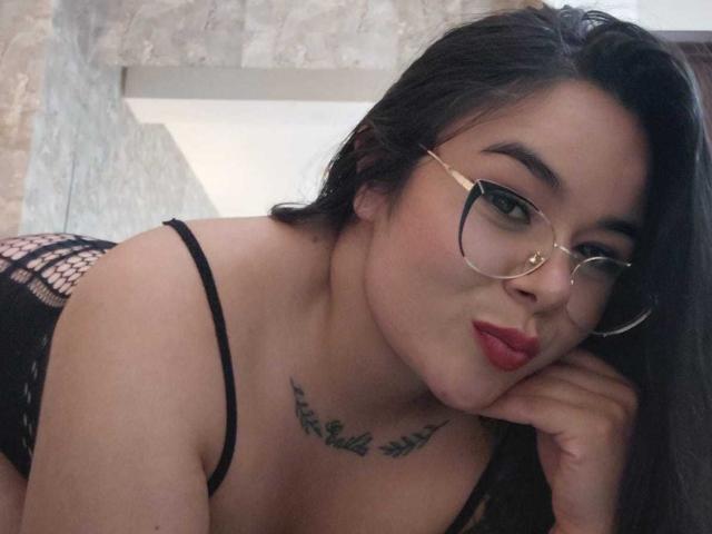 EveWillis with dark hair, glasses, red lipstick, black lace lingerie, and a tattoo under her tits, leaning forward, pouting.