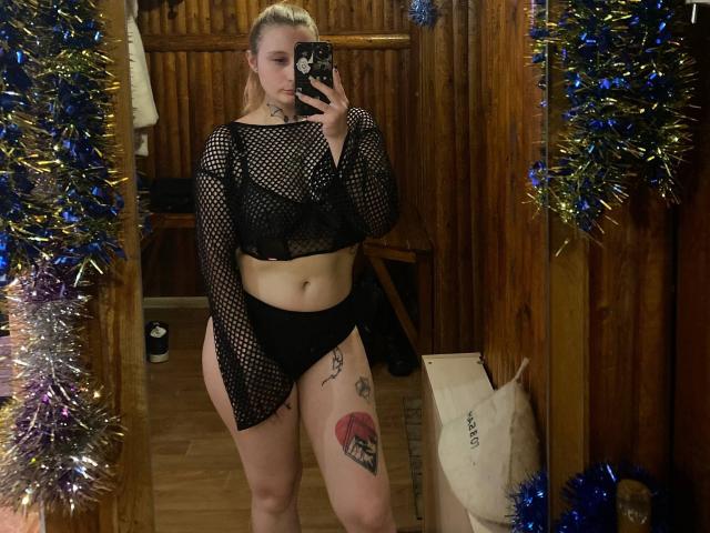CharlesettaLahr with a tattoo on her right thigh, wearing a black mesh top and black panties, holding a phone, standing in a wooden room with blue and silver tinsel.