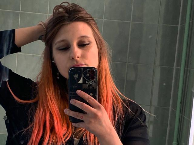 CharlesettaLahr with orange-dyed hair, black shirt, holding phone, eyes closed, hand in hair, mirror selfie, green-tiled wall background, serious expression, visible tattoo on wrist.