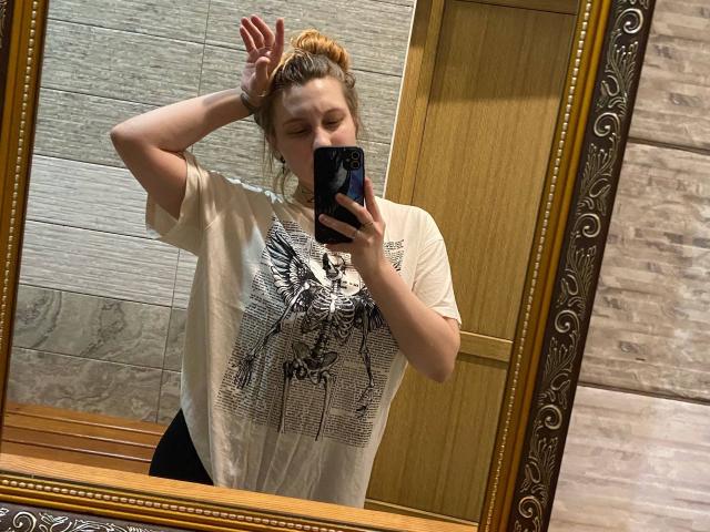 CharlesettaLahr with orange hair in a bun, wearing a white tee with a black graphic, taking a mirror selfie, right hand in hair, left hand holding phone.