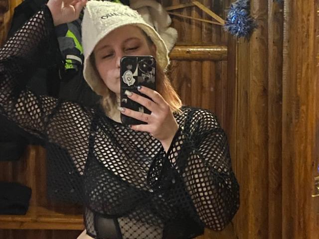 CharlesettaLahr with eyes closed, wearing a white knit hat and black fishnet top, showing her tits, taking a mirror selfie with a phone. Wooden wall background.