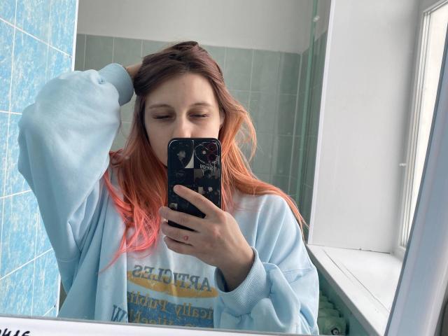 CharlesettaLahr with pink-dyed hair, wearing a light blue sweatshirt, taking a mirror selfie with her right hand in her hair, using a black phone with a dark case.