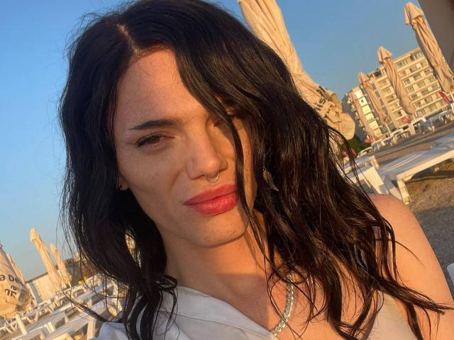 VanessaDomy with wet black hair, nose ring, and pink lipstick, wearing a white top with a silver necklace, sunlight on her face, looking slightly to the side.