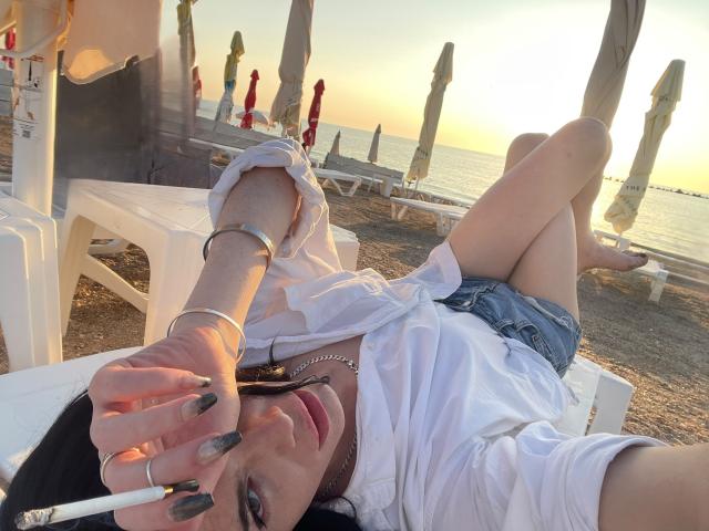 VanessaDomy ligt op een strandstoel, een sigaret rokend, gekleed in een wit shirt, spijkerbroekshorts en een ketting, met gekruiste benen, en met zonneparasols op de achtergrond..