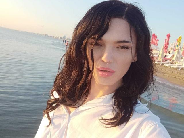 Vanessa, con i capelli castano scuro ondulati, una camicia bianca, labbra rosa e uno sguardo dolce, è in piedi vicino a una spiaggia tranquilla, con il sole che le riflette sul viso. In secondo piano, si vedono l'acqua e degli ombrelloni rossi..