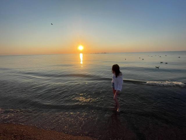 VanessaDomy, met haar achterkant naar de camera gericht, staand in ondiep water, gekleed in een witte jurk, kijkend naar de zonsondergang, vogels die in het water drijven, een zandstrand op de voorgrond, een kalme oceaan, een heldere lucht..