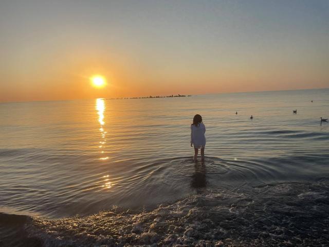 VanessaDomy staat in ondiep water bij zonsondergang, gekleed in een witte jurk, met haar rug naar de camera, en haar spiegelbeeld weerspiegelt in de kalme zee, omringd door kleine, verre, drijvende figuren..