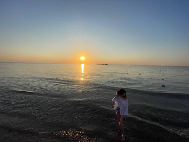 VanessaDomy in a white shirt standing in shallow water at sunset, sun reflection on water, small birds in water, calm ocean, soft evening light, gentle waves.