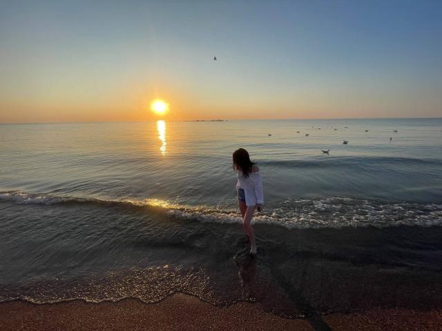 VanessaDomy with medium-length brown hair, wearing a white shirt, standing in shallow water at sunset, looking down at the water, small birds flying above, reflection on water.