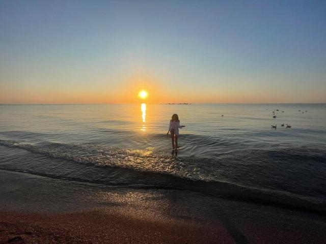 VanessaDomy staat in ondiep water, gekleed in een witte jurk, met de zon die op de oceaan reflecteert, kleine eenden die in de buurt zwemmen, en een zandstrand op de voorgrond..