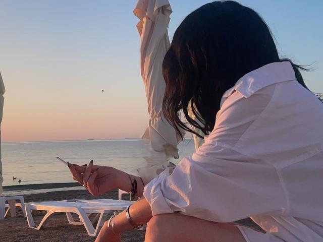 VanessaDomy, con cabello oscuro, vestida con una camisa blanca, agachada en la playa, sosteniendo un cigarrillo, una silla de playa blanca, atardecer, agua tranquila, un pequeño pájaro en el cielo..