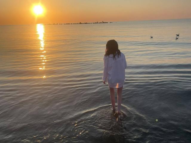 VanessaDomy met golvend bruin haar, gekleed in een witte blouse, staat in ondiep water, met haar gezicht naar de ondergaande zon gericht. Er is een reflectie van de zon op het wateroppervlak en vogels op de achtergrond..