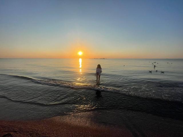 VanessaDomy stands in shallow water, back to the camera, at sunset. Her small ass reflects in the gentle waves. A glowing sun sets on the horizon. Birds float nearby.