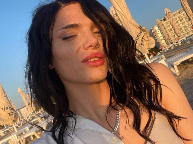 VanessaDomy with long black hair, nose ring, and pink lipstick, eyes closed, sunlit, white shirt, silver necklace, slight smile, soft light, beach umbrella, clear blue sky.
