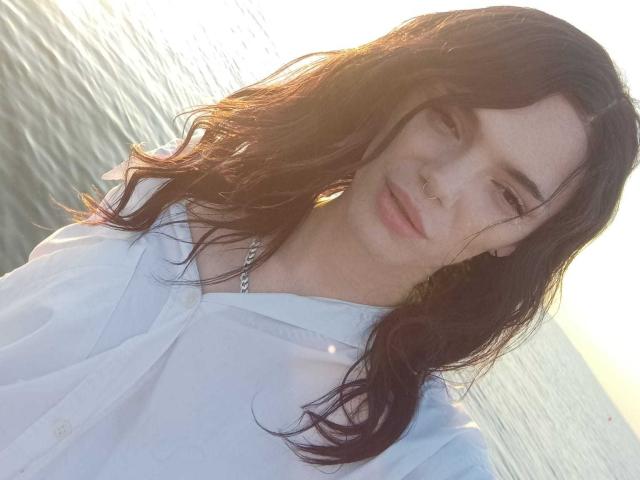 VanessaDomy met golvend bruin haar, een wit shirt, een neusring en een ketting, lachend naar de camera, met water en zon op de achtergrond..