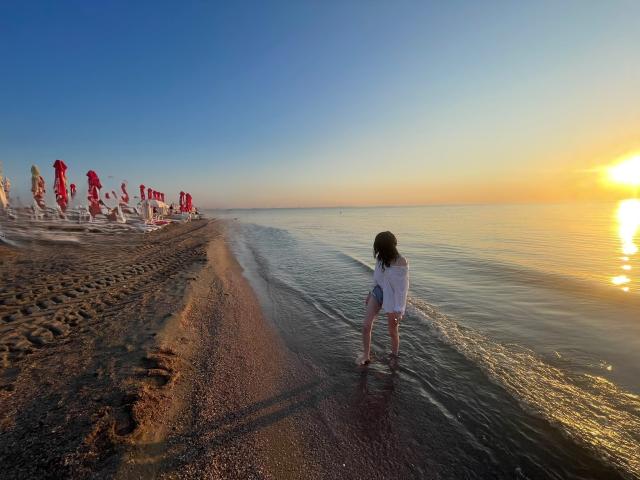 VanessaDomy stands in shallow water, wearing a white dress, with her back to the camera, sun setting on the right, red umbrellas on the left, sandy beach.