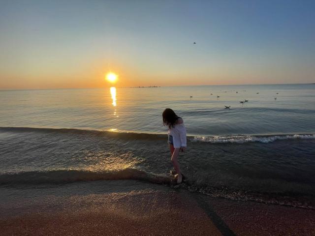 VanessaDomy avec une silhouette en robe blanche, debout près de l'eau au coucher du soleil, portant des baskets, regardant vers le bas, petites vagues, reflet sur l'eau, oiseaux volant, ciel clair, plage de sable..