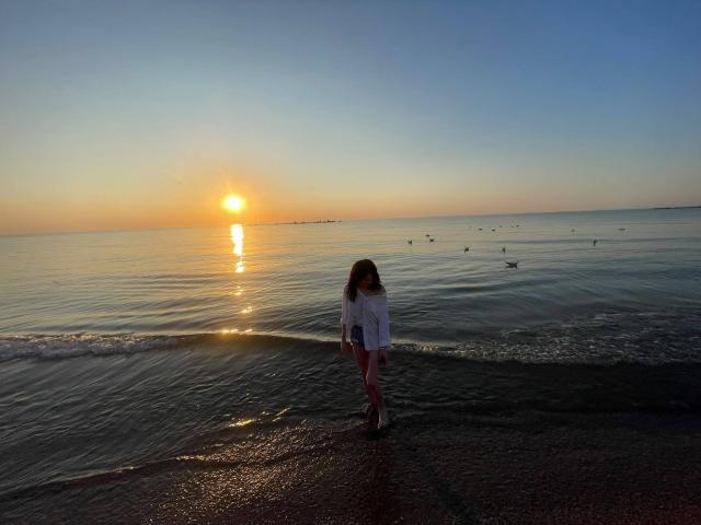 VanessaDomy in een witte blouse, staand aan de kust, de zonsondergang weerspiegelt in het water, kleine vogels vliegen rond, haar rug naar de camera gericht, haar achterkant is licht zichtbaar, nat zand..