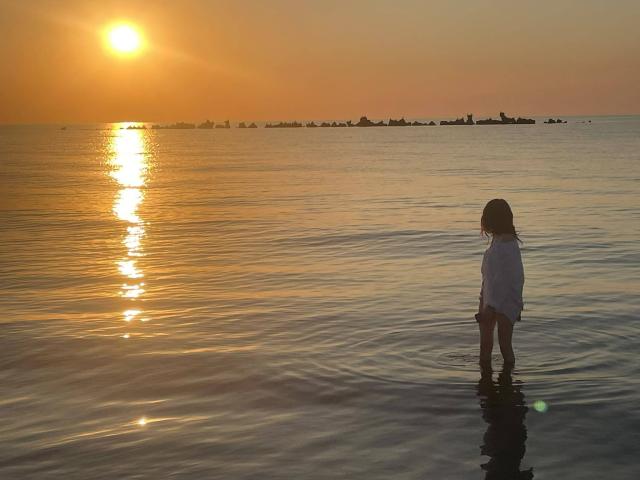 VanessaDomy staat in ondiep water bij zonsondergang, gekleed in een wit shirt, met haar achterkant nauwelijks boven het wateroppervlak, waarbij het gouden zonlicht op haar rug wordt weerkaatst..