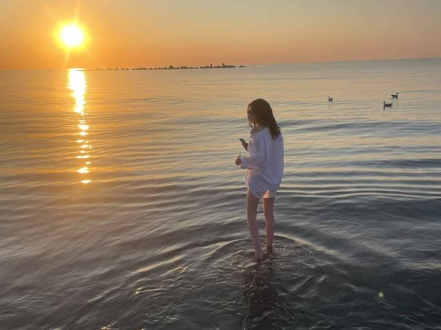 VanessaDomy staat in ondiep water, gekleed in een oversized wit shirt, en kijkt naar een telefoon. Achter haar is een zonsondergang te zien die reflecteert op de kalme oceaan, en er zijn kleine vogels op de achtergrond..