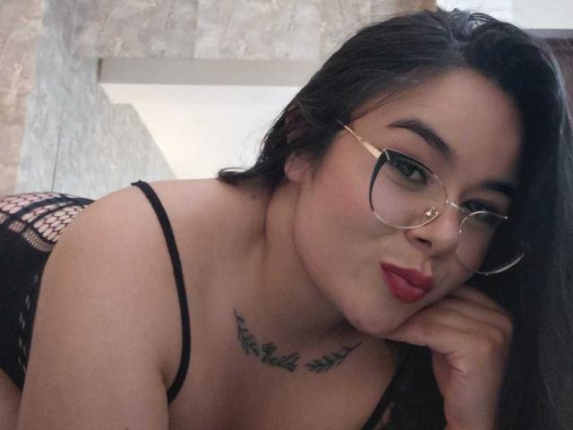EveWillis with glasses, black fishnet lingerie, pouty red lips, and a chest tattoo, leaning on her hand, showing her tits and ass in a sexy, confident pose.