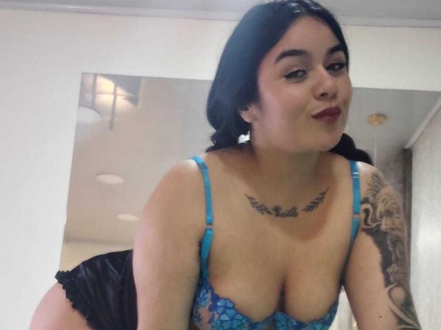 EveWillis with black hair, wearing a blue lace bra, black shorts, showing her large tits, tattoo on chest, tattoo on arm, smiling, looking at camera.