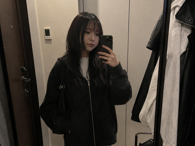 Annetta Hatchcock met lang zwart haar, gekleed in een zwarte hoodie met rits en een wit T-shirt, die een telefoon vasthoudt, een spiegelzelfportret, kijkt naar de camera, met een deur en kleding op de achtergrond..