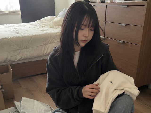 AnnettaHatchcock, ragazza asiatica con lunghi capelli neri, che indossa una felpa nera, tiene in mano un maglione bianco, seduta su un pavimento di legno, guarda in basso, camera da letto con letto e comò sullo sfondo..