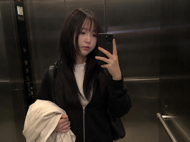 Annetta Hatchcock mit langen, dunklen Haaren, die einen schwarzen Mantel und ein weißes Hemd trägt, hält ein Telefon, ein weißes Handtuch in der linken Hand, wirkt ernst in einem Aufzug, silberne Wände..