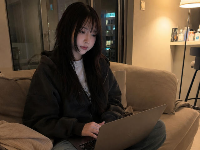 Annetta Hatchcock, com cabelos longos e pretos, vestindo um moletom escuro e uma camiseta branca, está sentada em um sofá bege, digitando em um laptop, em um quarto com pouca luz..
