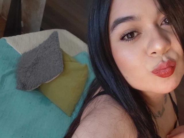 EveWillis with long black hair, red lipstick, and a slight pout, showing her face and a small tattoo on her chest, with colorful pillows in the background.